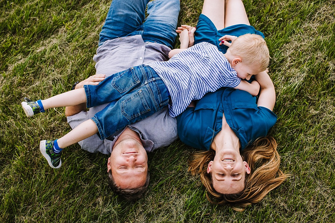 mama i tata leżą na plecach na trawie, a ich kilkuletni synek leży na nich w poprzek